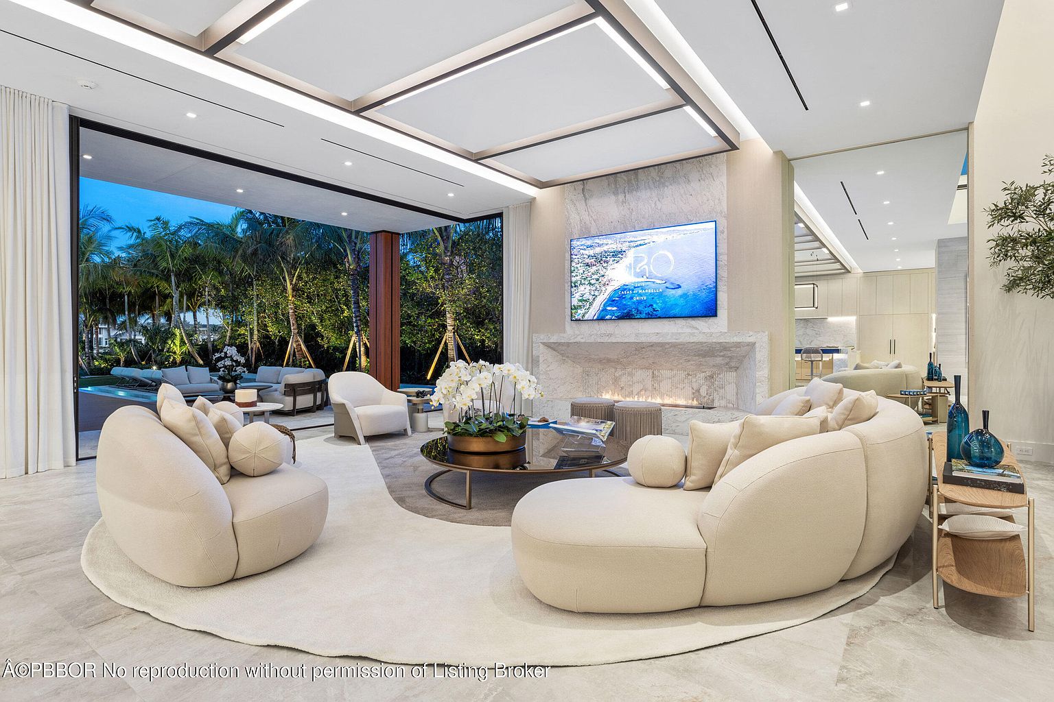 This is a luxurious living room featuring a modern design with neutral tones and high-end finishes. The room includes a large curved sofa, a round coffee table, and a fireplace with a TV mounted above. Large sliding glass doors open to an outdoor area, creating a seamless indoor-outdoor living experience.