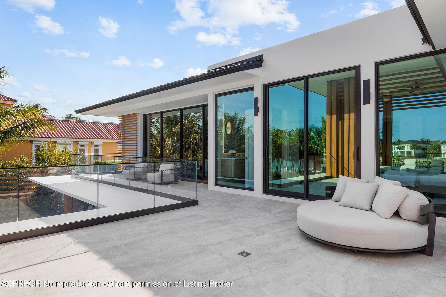 This image showcases a luxurious outdoor patio or balcony area. The space features modern design elements, including sleek glass railings, contemporary outdoor furniture, and large sliding glass doors that seamlessly blend indoor and outdoor living. The patio is finished with light-colored stone tiles, creating a bright and inviting atmosphere.