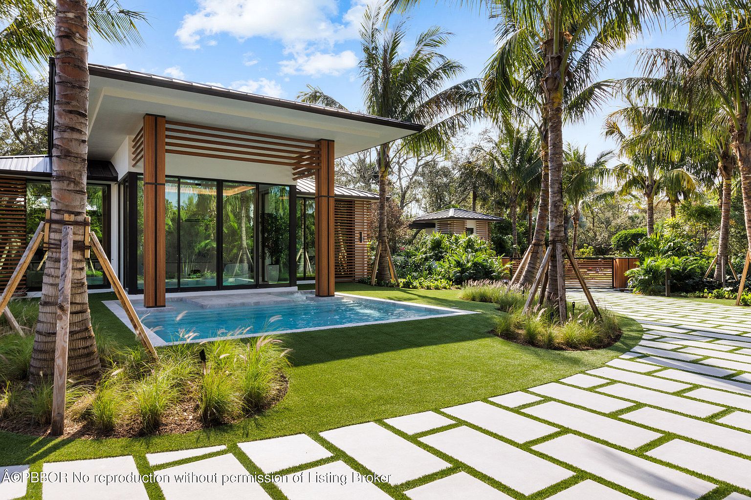 This image showcases a modern outdoor pool and spa area, featuring a sleek, contemporary building with large glass windows and wooden support beams. The pool is surrounded by manicured green grass and palm trees, creating a tropical oasis. A paved walkway with grass in between the stones leads to the pool area, enhancing the property's curb appeal and luxurious feel.