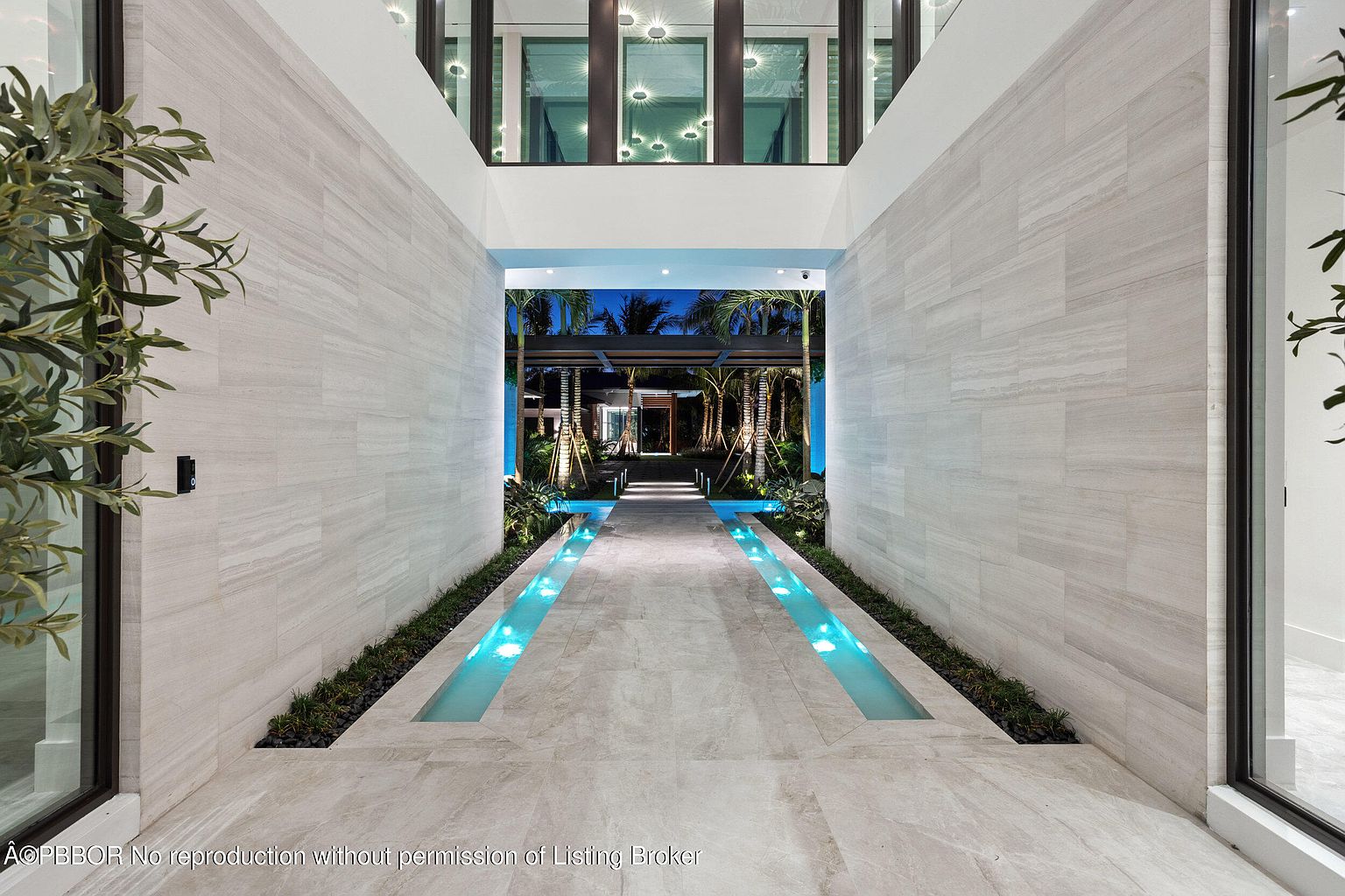 This image showcases a grand entryway featuring a long, tiled walkway with integrated water features on either side, illuminated with blue lighting. The walkway is flanked by high, textured walls, leading to an open outdoor area with palm trees and a covered structure. The perspective is a straight-on view, emphasizing the depth and architectural design of the entrance.
