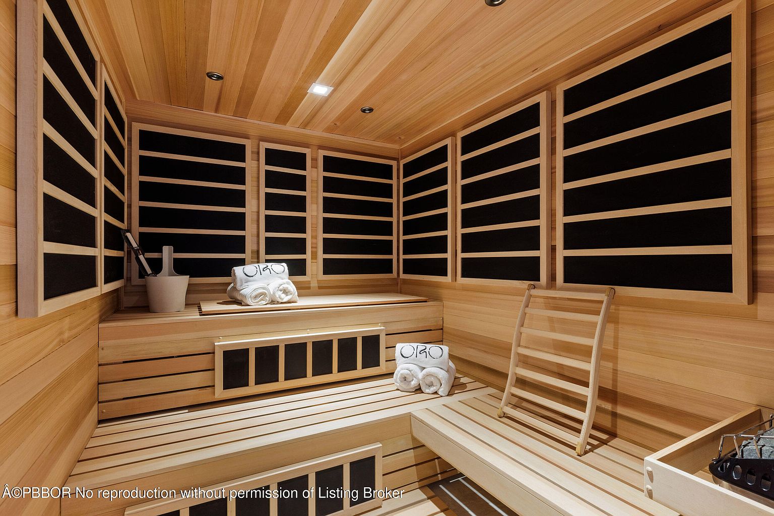 This is an interior shot of a sauna, featuring light-colored wood paneling on the walls, ceiling, and benches. The sauna includes tiered seating, black panel heaters, and accessories such as towels and a bucket with a ladle. The overall impression is clean, modern, and relaxing, suggesting a high-end spa-like experience.