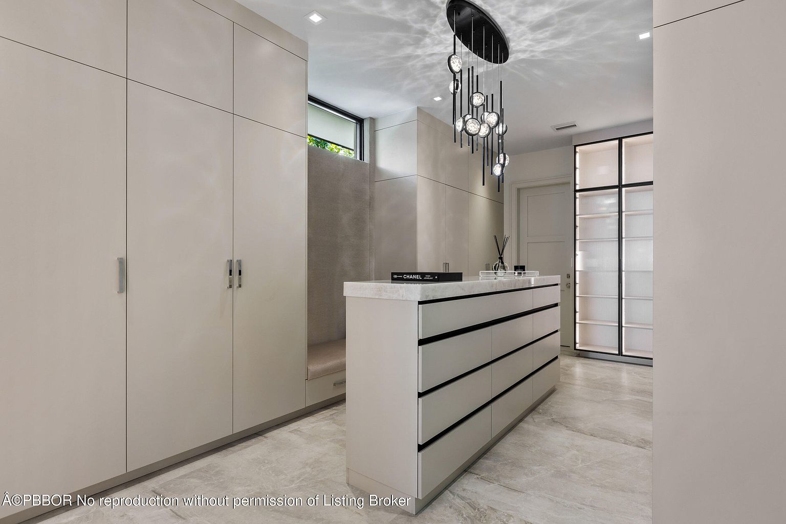 This is a well-organized walk-in closet featuring custom cabinetry in a neutral color palette. The closet includes ample storage with drawers and shelving, and a stylish chandelier adds a touch of elegance. The flooring appears to be marble or a similar stone, contributing to the luxurious feel of the space.
