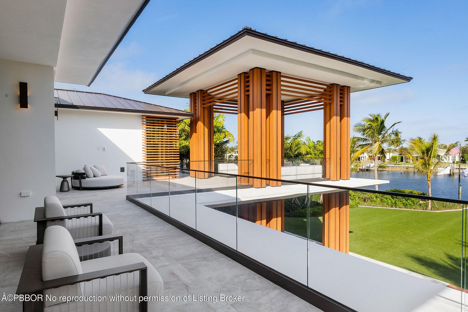 This image showcases a luxurious outdoor patio/deck/balcony area with modern architectural design. The space features sleek furniture, a glass railing offering unobstructed views of the waterfront, and a unique wooden structure providing shade and visual interest. The overall impression is one of sophisticated outdoor living and relaxation.