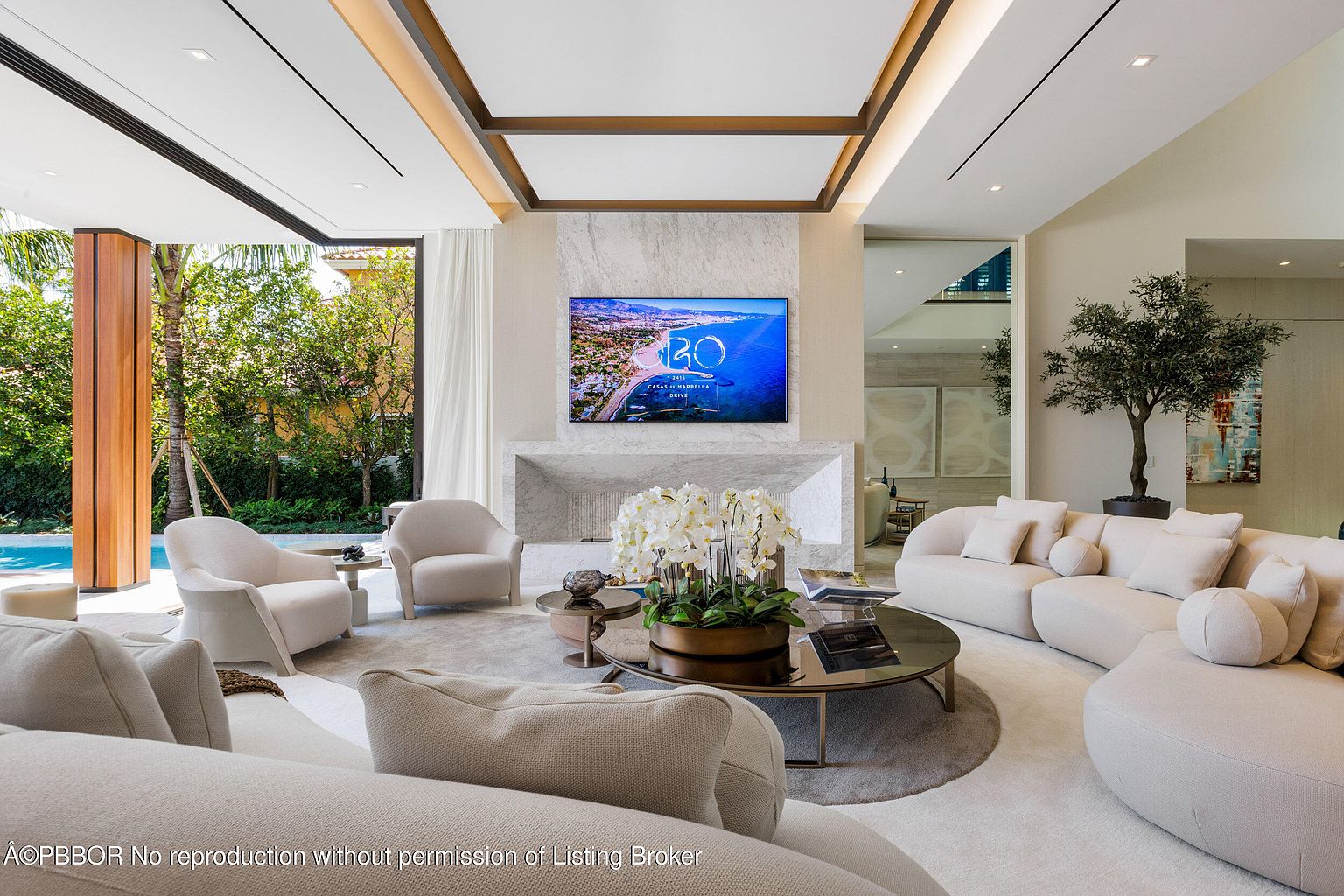 This is a luxurious living room featuring a neutral color palette with white sofas and armchairs arranged around a round coffee table. A large television is mounted above a marble fireplace, and the room is filled with natural light from the adjacent outdoor area. An olive tree in a pot adds a touch of greenery to the space.