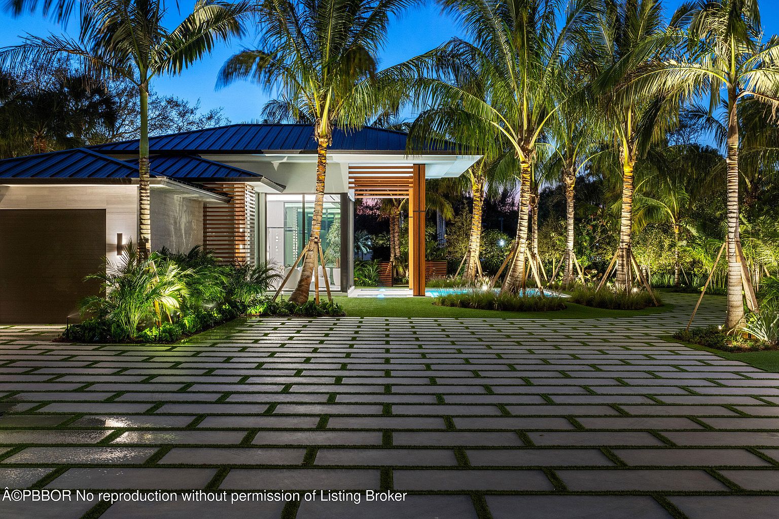 This image showcases the front exterior of a modern home at dusk, featuring a meticulously designed driveway with grass accents between the pavers. The property is adorned with several palm trees, strategically lit to highlight their architectural beauty. The home's facade includes a combination of materials, such as wood and stone, creating a sophisticated and inviting entrance.