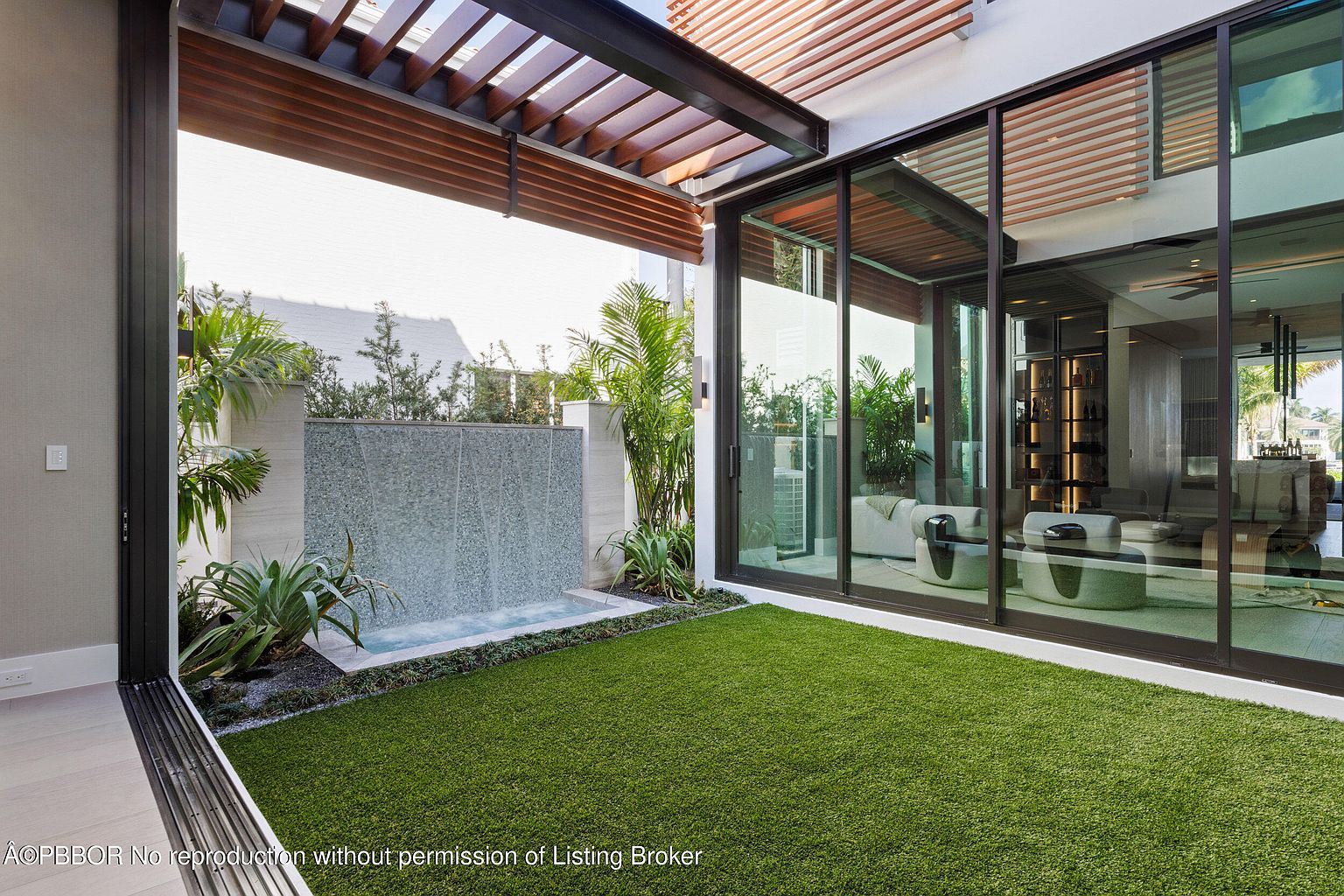 This image showcases a modern patio area with a sleek design. A water feature adds a tranquil element, complemented by artificial turf for a low-maintenance green space. Large glass doors provide seamless indoor-outdoor living, offering a glimpse into the home's interior.