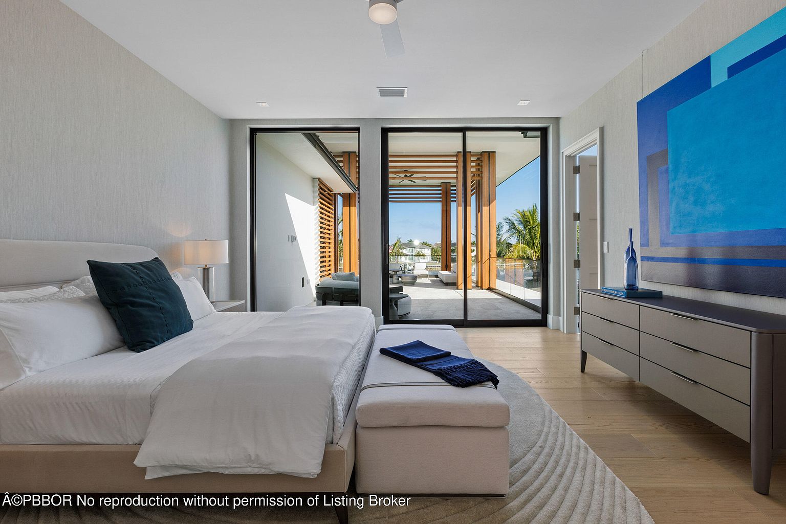 This is a bright and airy primary bedroom featuring a large bed with white linens and a dark blue accent pillow. Sliding glass doors lead to an outdoor balcony, offering natural light and a view. A modern dresser with a blue abstract painting above it adds a touch of color and style to the room.