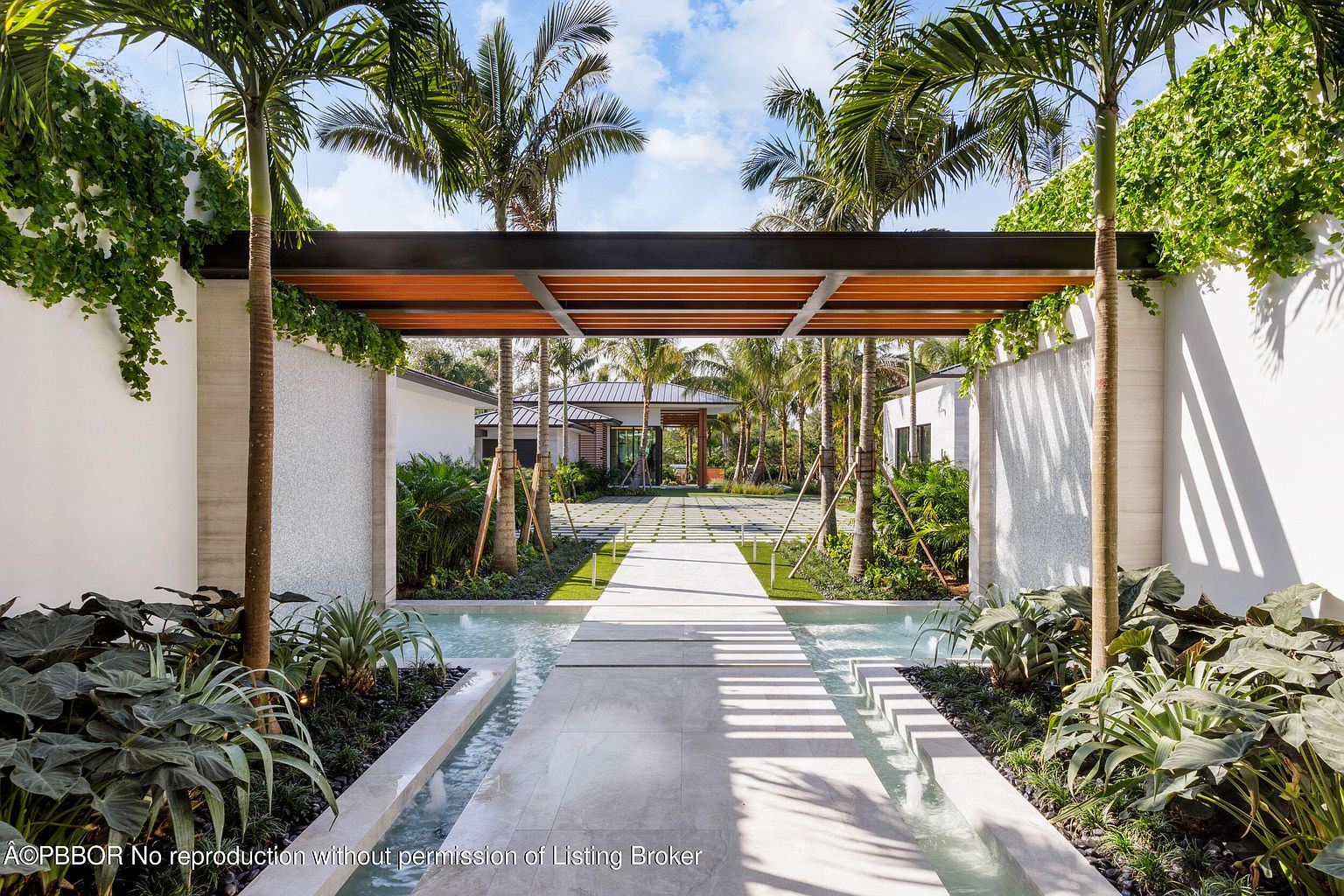 This image showcases a stunning entryway featuring a modern pergola structure with wooden slats and a dark metal frame, creating a shaded walkway. A shallow water feature runs along the sides of the pathway, adding a serene and luxurious touch. Lush tropical landscaping and palm trees frame the view, leading to a glimpse of the house beyond, creating an inviting and sophisticated first impression.