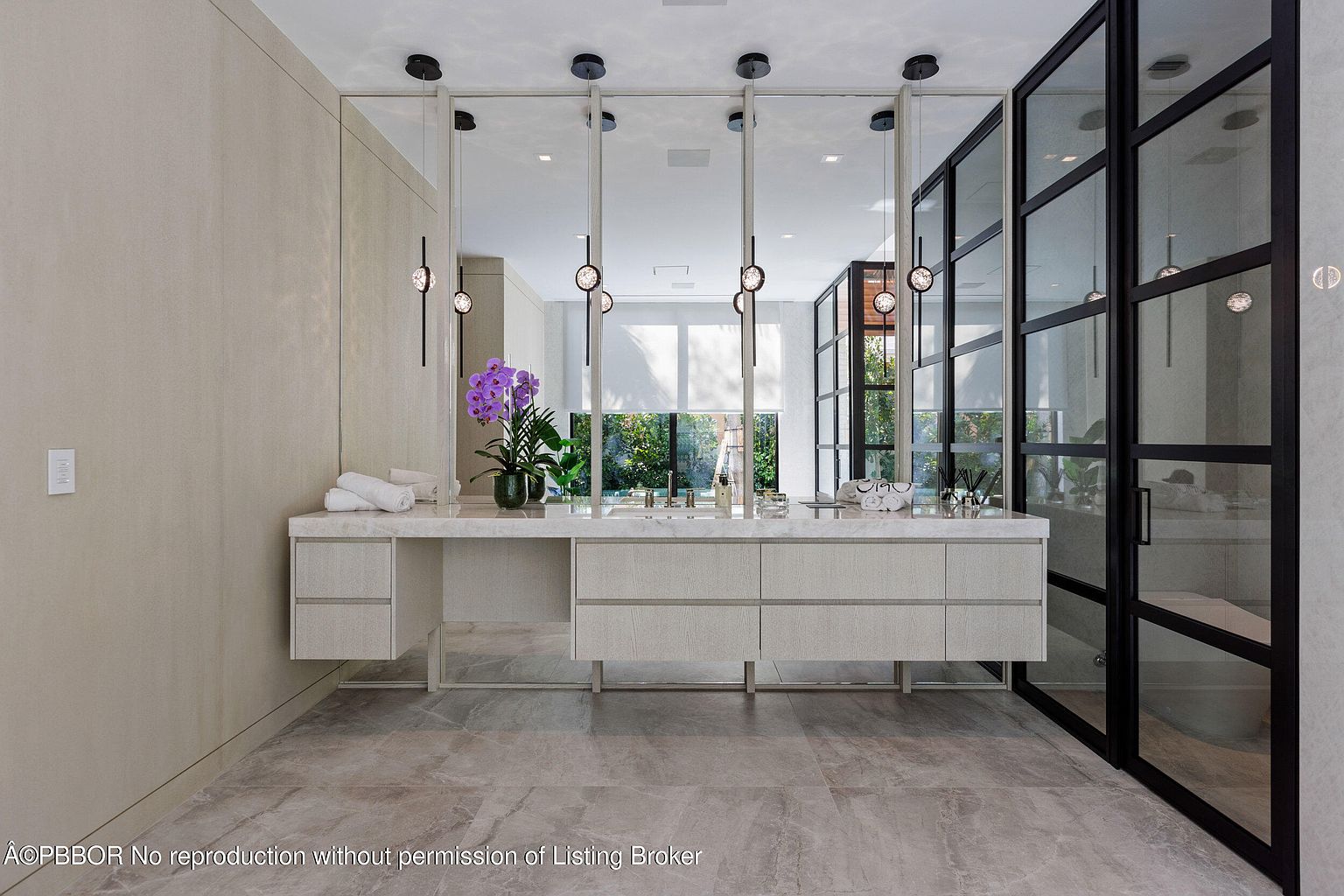 This is a luxurious primary bathroom featuring a long, modern vanity with multiple drawers and a marble countertop. The vanity is illuminated by pendant lights hanging from the ceiling, and a large mirror spans the length of the vanity. A glass-enclosed shower is visible on the right, and the flooring is a light-colored tile. The overall impression is clean, elegant, and spa-like.