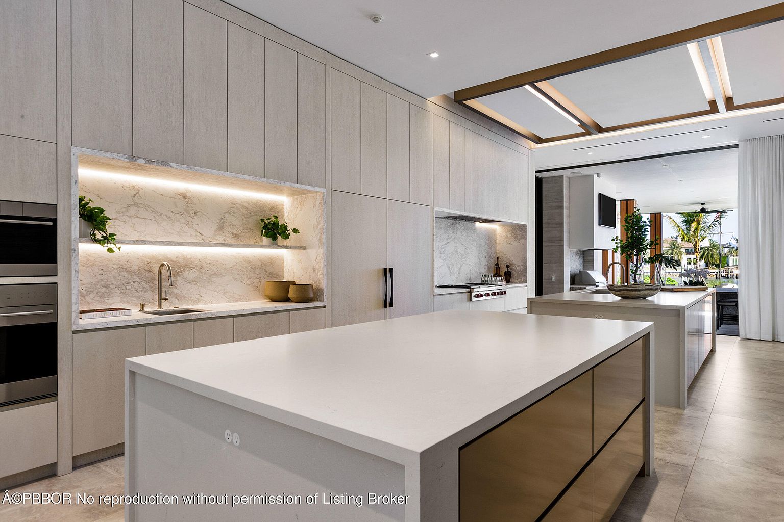 This is a bright and modern kitchen featuring sleek, light-colored cabinetry and a large island with a white countertop. The kitchen includes built-in appliances and a marble backsplash, complemented by recessed lighting and a unique ceiling design. The open layout leads to an outdoor view, enhancing the sense of space and luxury.