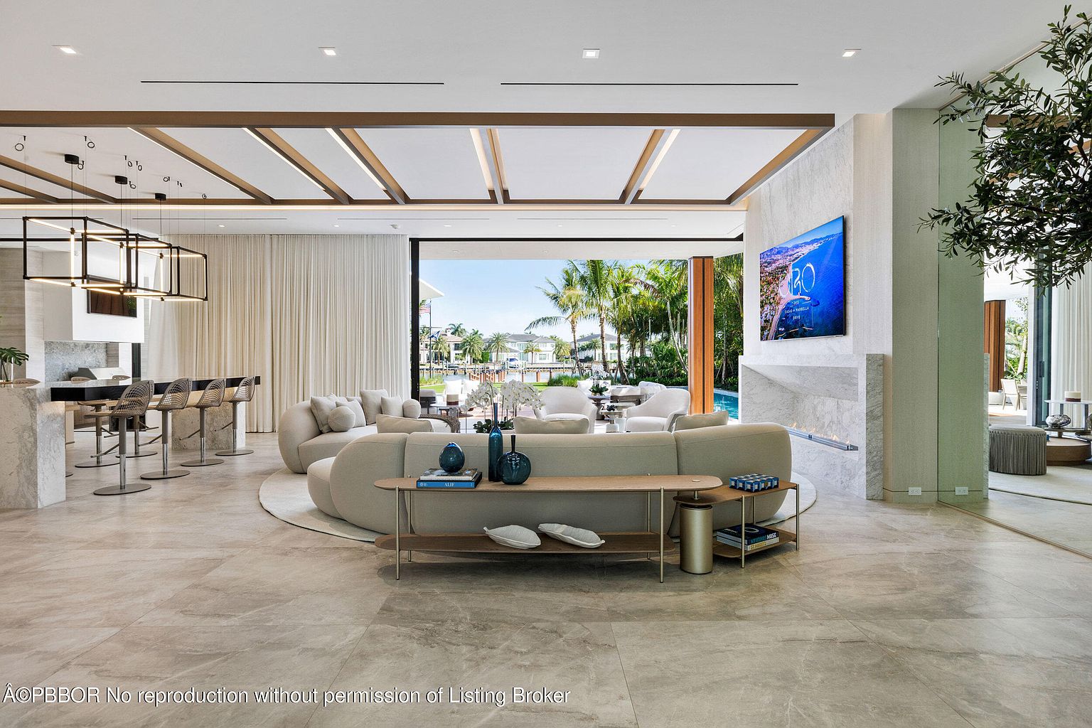 This is a luxurious living room featuring a large, curved sofa, a modern entertainment center with a flat-screen TV, and an open floor plan that leads to an outdoor area with a pool. The room is decorated in neutral tones with pops of blue, and the flooring is a light-colored tile. The overall impression is one of spaciousness and high-end design.