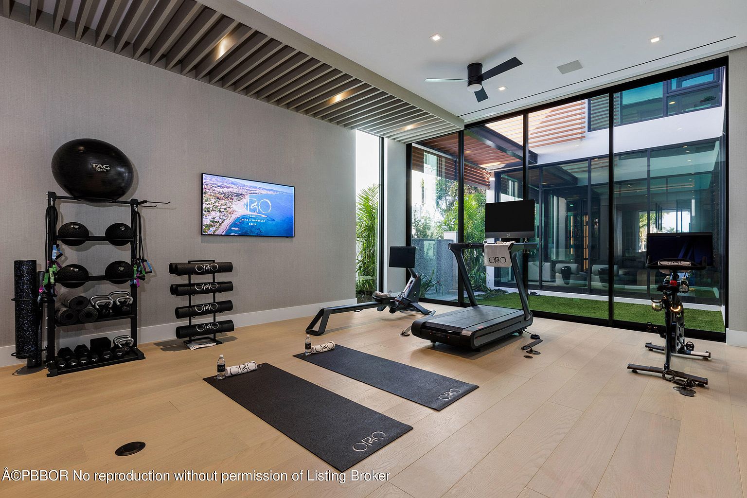 This is a well-equipped home gym featuring a variety of exercise equipment including a treadmill, stationary bike, rowing machine, and free weights. The room has a modern aesthetic with light wood flooring, a large flat-screen TV, and floor-to-ceiling windows that offer a view of the exterior. The space is designed to be both functional and visually appealing, creating an inviting workout environment.