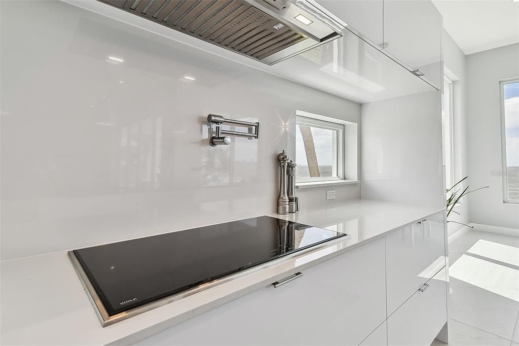 A sleek, modern kitchen features glossy white cabinetry and countertops, complemented by a black induction cooktop. A stainless steel pot filler is mounted on the backsplash, and a minimalist range hood is installed above the cooktop. The overall impression is clean, bright, and high-end.