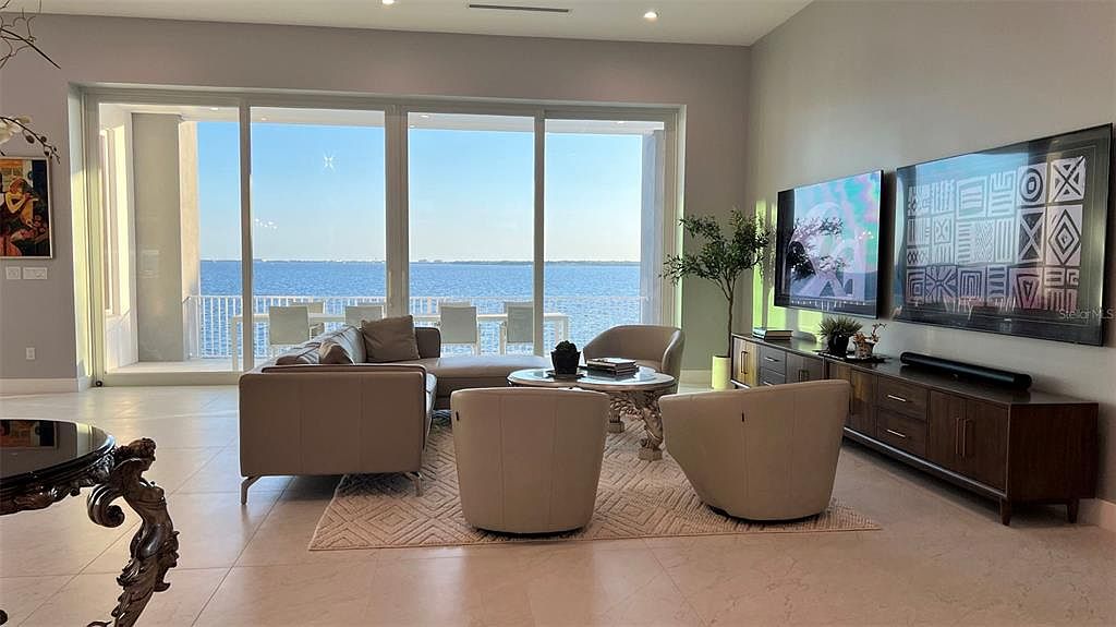 This is an interior shot of a living room with a modern design. The room features a large window with a view of the water, a sectional sofa, two armchairs, a round coffee table, and a media console with a television. The color palette is neutral, creating a serene and inviting atmosphere.