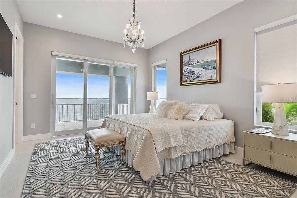 This is a primary bedroom featuring a bed with white linens, a decorative chandelier, and a framed painting on the wall. Sliding glass doors lead to a balcony with a water view, enhancing the room's appeal. The room is decorated in neutral tones with a patterned rug, creating a serene and luxurious atmosphere.