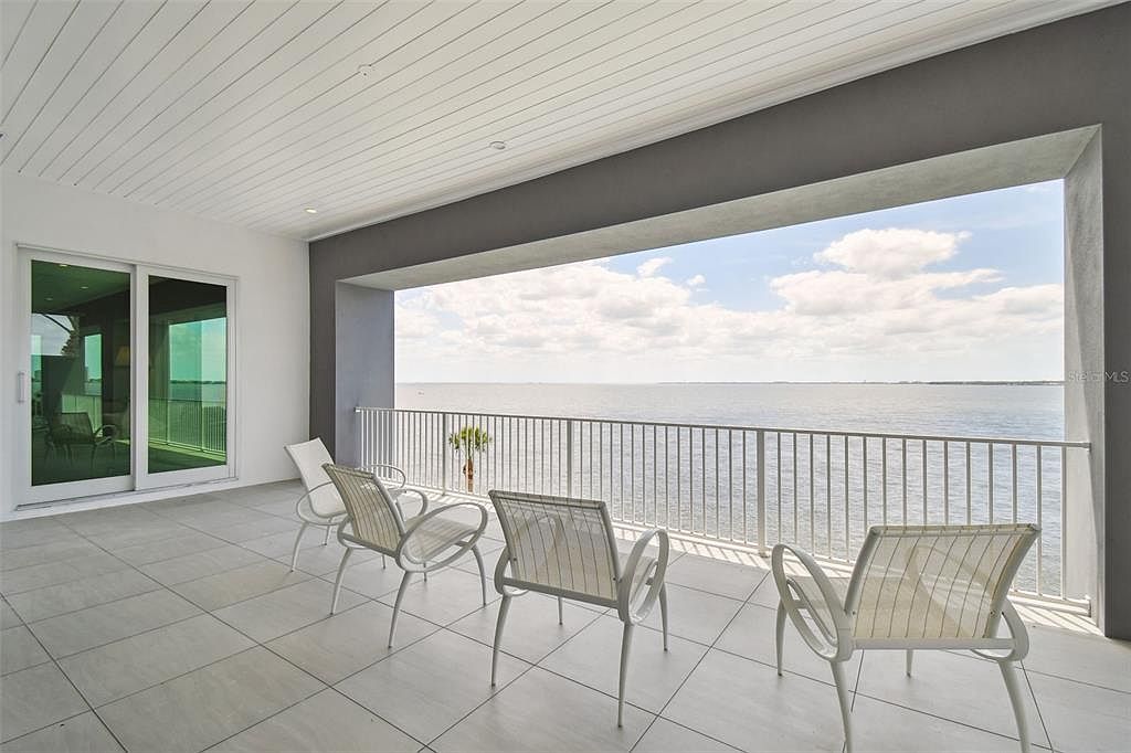 This image showcases a spacious balcony with a stunning waterfront view. The balcony features modern white chairs, light gray tiled flooring, and a sleek railing. A large sliding glass door provides access to the interior, while the open design maximizes the scenic vista, creating an inviting outdoor living space.