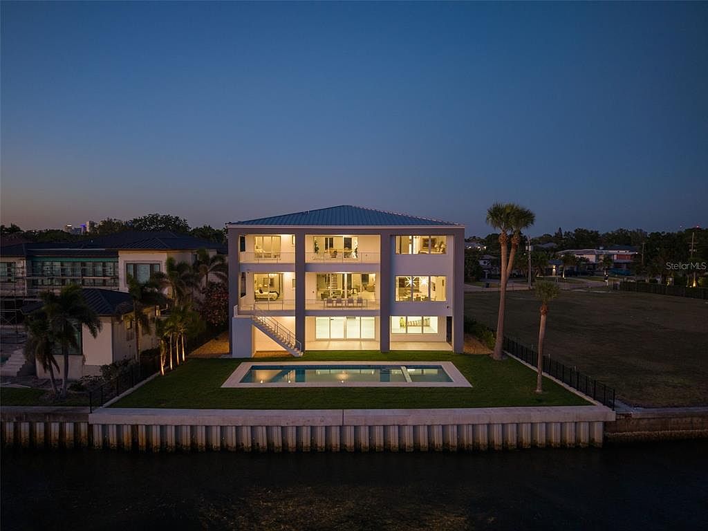 This is a rear exterior view of a modern, multi-story waterfront home at dusk. The house features large windows and balconies, offering ample natural light and outdoor living space. A rectangular pool is situated in the backyard, which is neatly landscaped with grass and palm trees, and a seawall protects the property from the water.