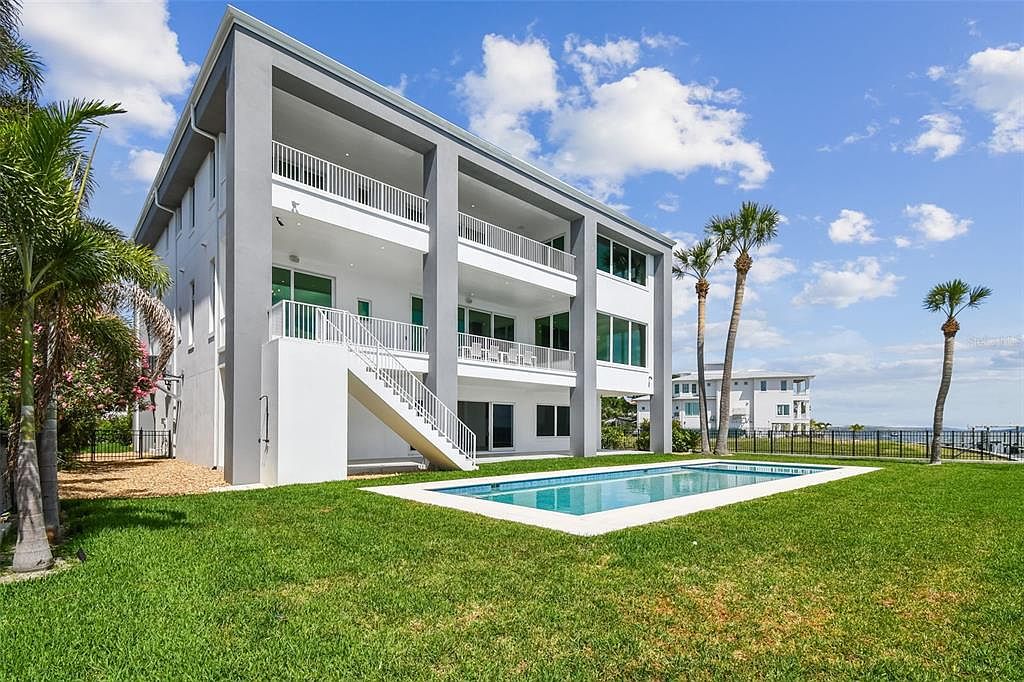 This image showcases the rear exterior of a modern, multi-story home, emphasizing its outdoor living space. A rectangular pool with a light-colored border is centered in the well-manicured lawn, complemented by palm trees and a glimpse of the waterfront in the background. The home features multiple balconies with white railings, adding to its luxurious appeal.