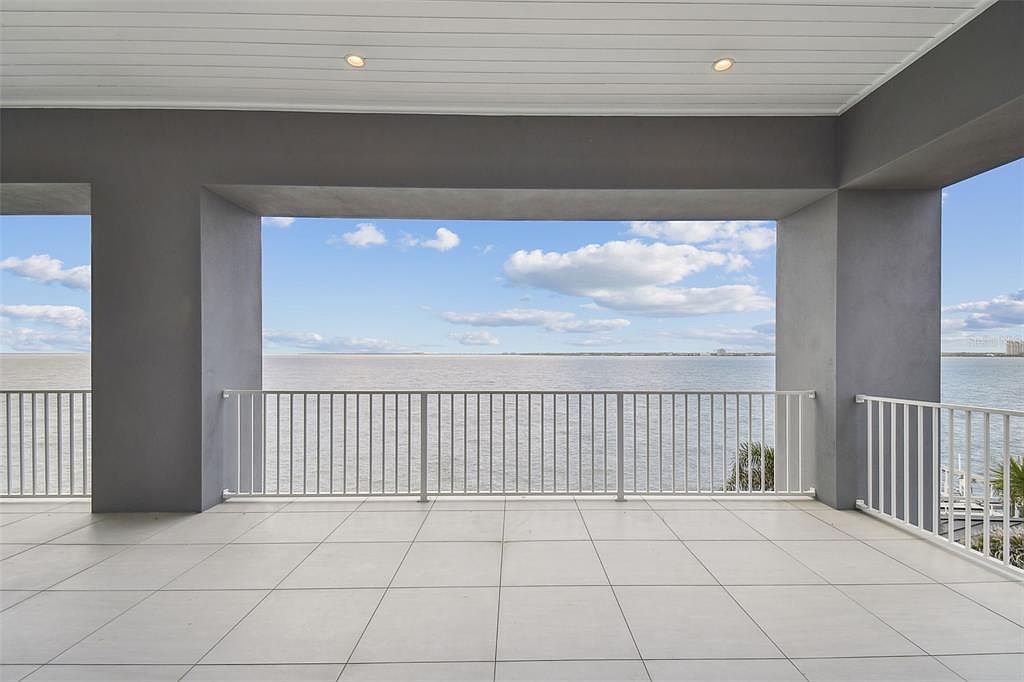 This image showcases a spacious balcony with a tiled floor and a white railing, offering an expansive view of the ocean under a partly cloudy sky. The balcony is framed by gray structural columns and a ceiling with recessed lighting, creating a luxurious and serene outdoor living space. The perspective is from inside the balcony looking out towards the ocean.