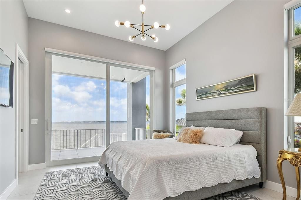 This is a bright and airy primary bedroom featuring a large sliding glass door with a view of the water. The room is decorated in neutral tones with a gray upholstered bed frame, a white quilt, and a modern chandelier. The overall impression is luxurious and serene.