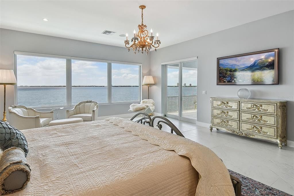 This is a luxurious primary bedroom featuring a large bed with a decorative headboard and a view of the water through large windows. The room is decorated in neutral tones with a chandelier and a dresser with a television above it. The overall impression is one of elegance and comfort.