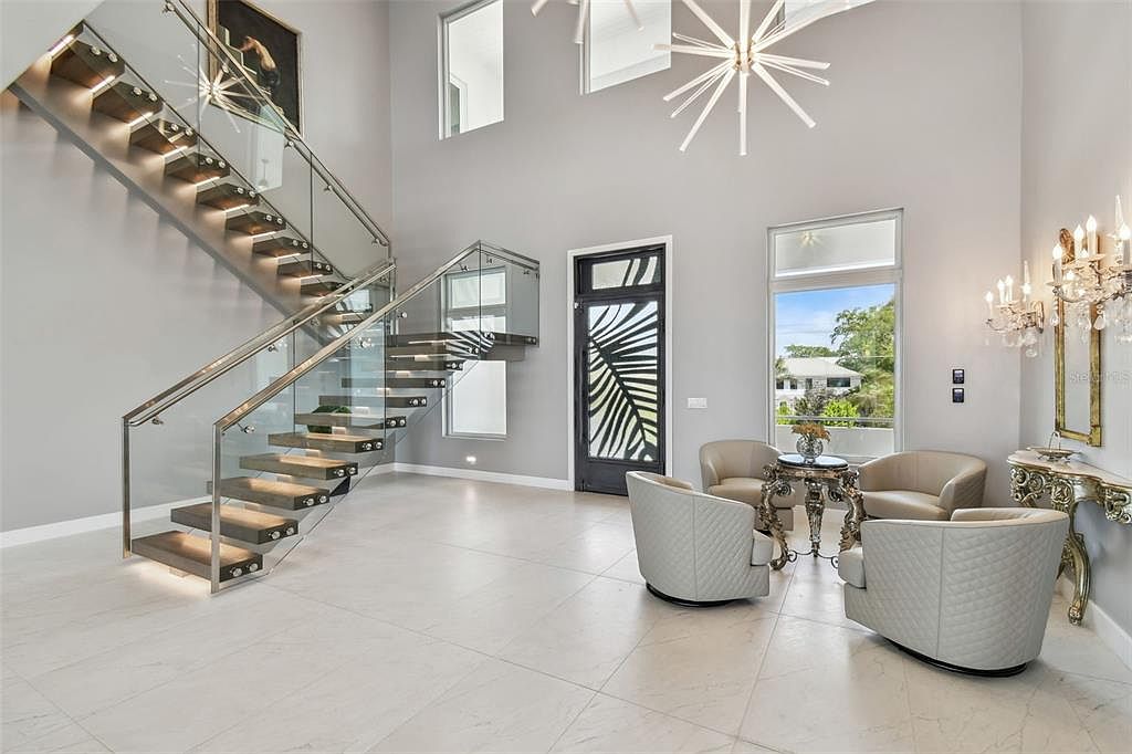 This is an interior shot of a grand foyer featuring a modern staircase with glass railings and floating wooden steps. The space is well-lit with a contemporary chandelier and natural light from nearby windows, complemented by elegant seating and a decorative door, creating a luxurious and inviting atmosphere.