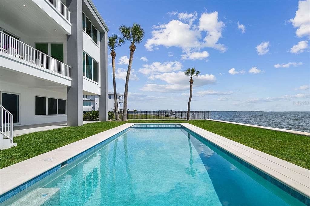 This image showcases a stunning rectangular swimming pool situated adjacent to a modern, multi-story home. The pool's clear blue water reflects the sky, and it is bordered by a well-maintained lawn and palm trees, creating a serene and luxurious outdoor space. The view extends to the ocean, enhancing the property's appeal and offering a sense of tranquility and exclusivity.
