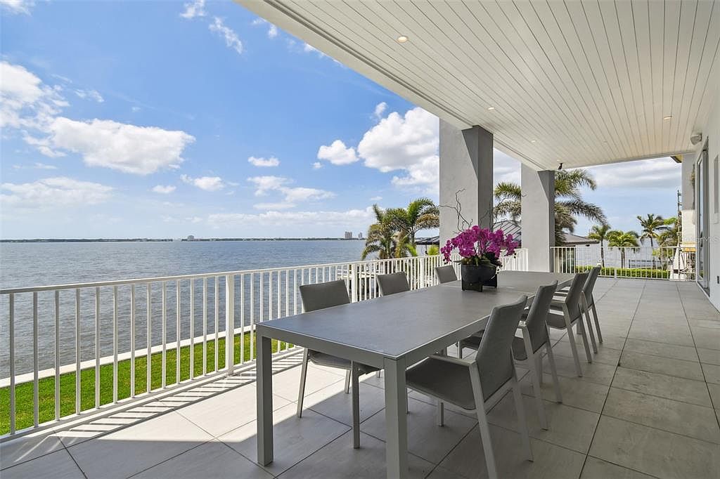 This image showcases a spacious outdoor patio or balcony area, featuring a large dining table with modern chairs, perfect for entertaining. The area is surrounded by a white railing, offering stunning views of the water and distant cityscape. The tiled flooring and covered ceiling add to the luxurious feel of this outdoor living space.