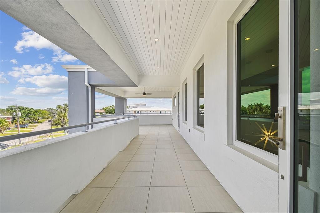 This image showcases a spacious outdoor balcony area with a modern design. The balcony features light-colored tile flooring, a white railing, and a covered ceiling with recessed lighting. Large windows and a glass door provide access to the interior, while offering views of the surrounding neighborhood and sky.