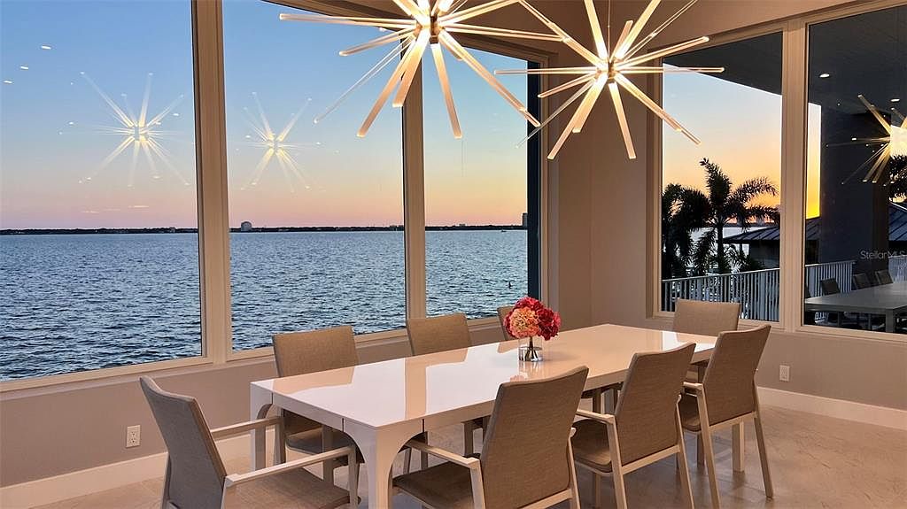 This is an interior shot of a dining room featuring a large white dining table with seating for eight. The room is well-lit by modern starburst chandeliers and natural light streaming through large windows that offer a scenic view of the water and sunset. The overall impression is elegant and serene, highlighting the room's spaciousness and connection to the outdoors.