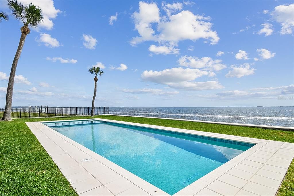This image showcases a rectangular swimming pool with a white tile surround, situated on a lush green lawn with a view of the ocean. The scene is set under a bright blue sky with scattered clouds, and palm trees add a tropical touch. A black metal fence is visible in the background, providing a sense of enclosure and safety.
