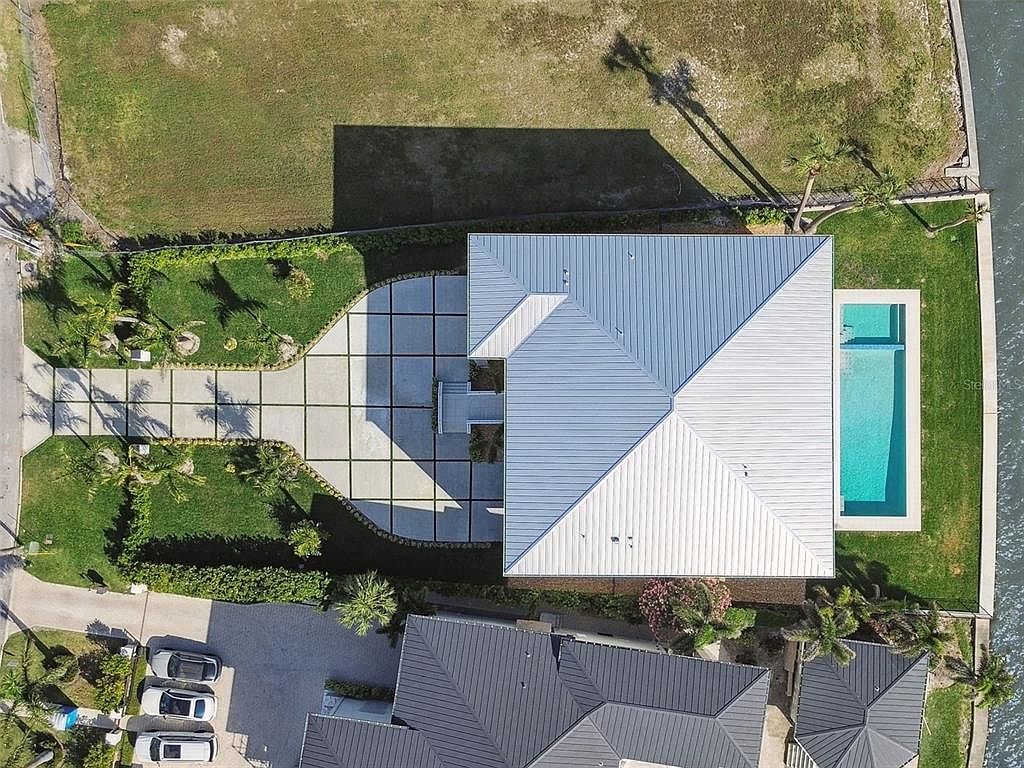 This aerial shot showcases a modern home with a light-colored metal roof, a rectangular swimming pool, and a meticulously designed driveway with geometric concrete pavers. The property features lush green lawns and landscaping, offering a clean and contemporary aesthetic. The home is situated near a canal, adding to its waterfront appeal.