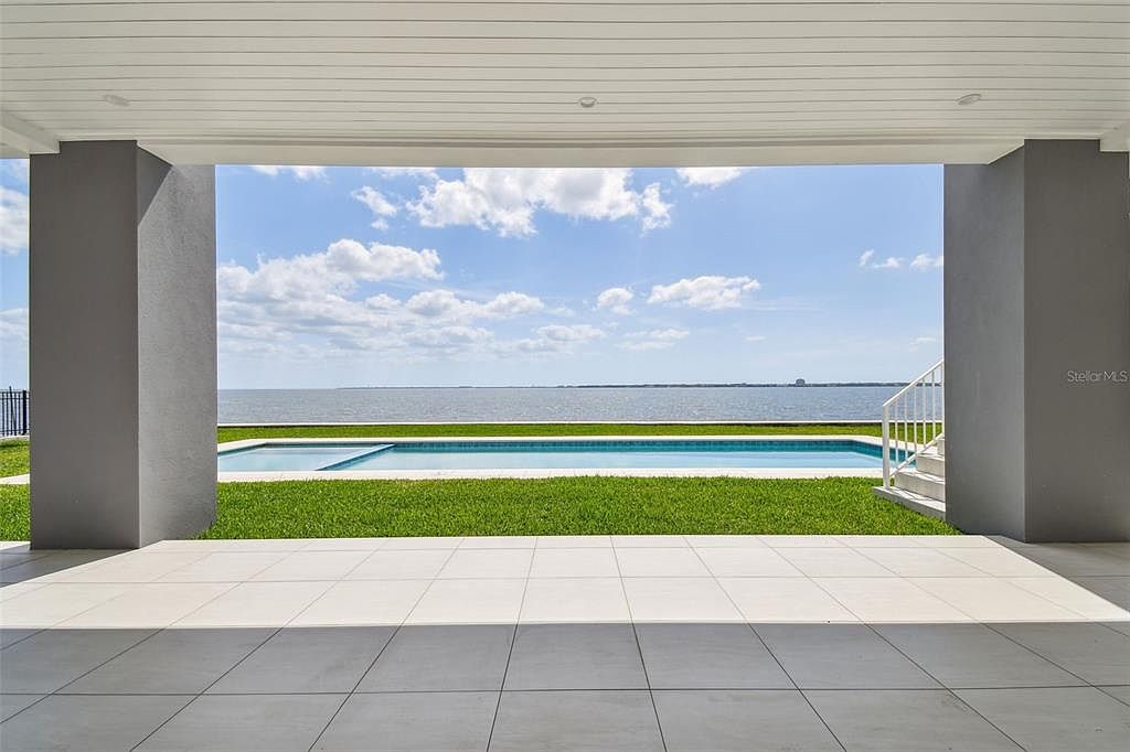 This image showcases a covered patio or balcony area with a view of a pool, lawn, and waterfront. The patio features light-colored tiled flooring and substantial gray support columns, creating a modern and open-air living space. The scene offers a seamless transition between indoor and outdoor living, emphasizing the property's waterfront location and recreational amenities.