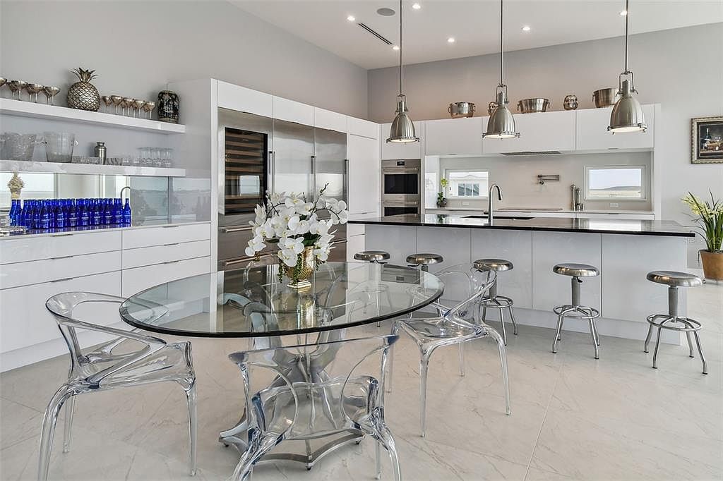This is a bright, modern kitchen featuring sleek white cabinetry, stainless steel appliances, and a black countertop island with bar seating. A glass-topped dining table with clear acrylic chairs sits in the foreground, complemented by pendant lighting above the island. The overall impression is clean, contemporary, and luxurious, with an open layout.