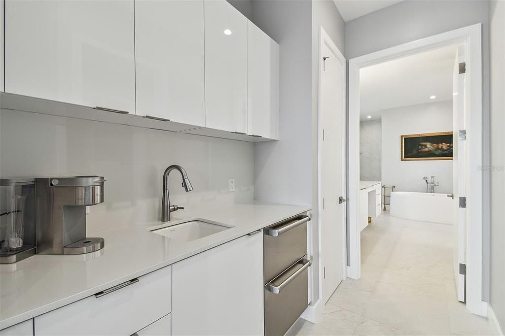 This is a bright, modern kitchen featuring sleek white cabinetry and countertops, complemented by stainless steel appliances. The kitchen area includes a sink and a beverage center, with an open doorway leading into a well-lit bathroom, creating a sense of flow and luxury. The perspective is from a medium shot, highlighting the kitchen's functionality and connection to the adjacent space.