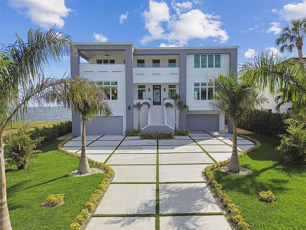 This is a front exterior view of a modern two-story home. The house features a symmetrical design with a white facade, gray accents, and large windows. A concrete driveway with grass inlays leads to the two-car garage and a set of stairs leading to the front door, flanked by palm trees and manicured landscaping.