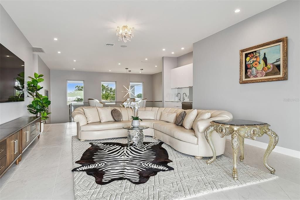 This is an interior shot of a living room featuring a large, cream-colored sectional sofa with decorative pillows, a zebra-print rug layered over a geometric-patterned area rug, and a glass-topped coffee table. A decorative side table with ornate legs sits beside the sofa, and a painting hangs on the wall. The room is well-lit with natural light from windows and recessed lighting, creating a bright and inviting atmosphere.