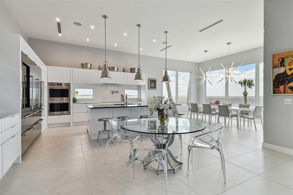 This is a bright and modern kitchen and dining area featuring sleek white cabinetry, stainless steel appliances, and a large island with a black countertop. A glass dining table with transparent chairs sits in the foreground, illuminated by contemporary pendant lights. Large windows offer a stunning view of the water, enhancing the open and airy feel of the space.