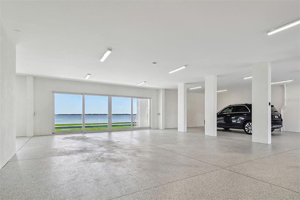 This is a spacious, modern garage with a view of the water. The garage features a polished concrete floor, white walls and ceiling, and large sliding glass doors that open to the outside. A black SUV is parked inside, and the space is well-lit with recessed lighting.