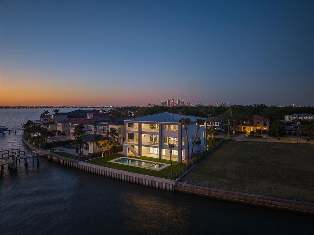 This aerial view showcases a modern waterfront home at dusk, featuring a clean, white exterior, a pool, and well-manicured landscaping. The property is situated on a canal with a seawall, offering direct water access. The city skyline is visible in the distance, adding to the property's appeal.