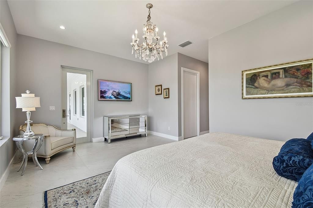 This is a primary bedroom featuring a chandelier, a mirrored dresser with a TV above it, and a large bed with blue pillows. The walls are painted a neutral gray, and there is artwork on the walls. The room has a luxurious and elegant feel.