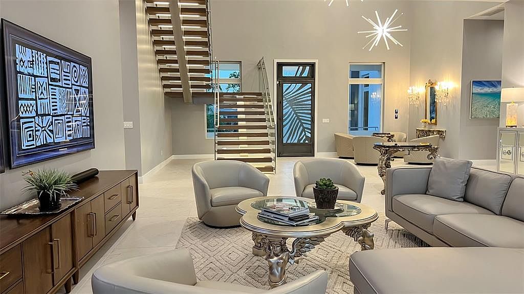 This is an interior shot of a modern living room featuring a floating staircase, a large television with abstract art, and contemporary furniture including a gray sofa and swivel chairs. The room is well-lit with a combination of natural light and modern light fixtures, creating a bright and inviting atmosphere. The perspective is from the front of the room, showcasing the open layout and stylish design elements.