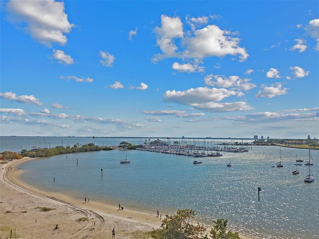 This aerial view showcases a beautiful coastal scene with a sandy beach curving around a calm bay. A marina filled with numerous boats is visible, and the clear blue sky is dotted with fluffy white clouds. The overall impression is serene and inviting, highlighting the property's proximity to the water and recreational opportunities.