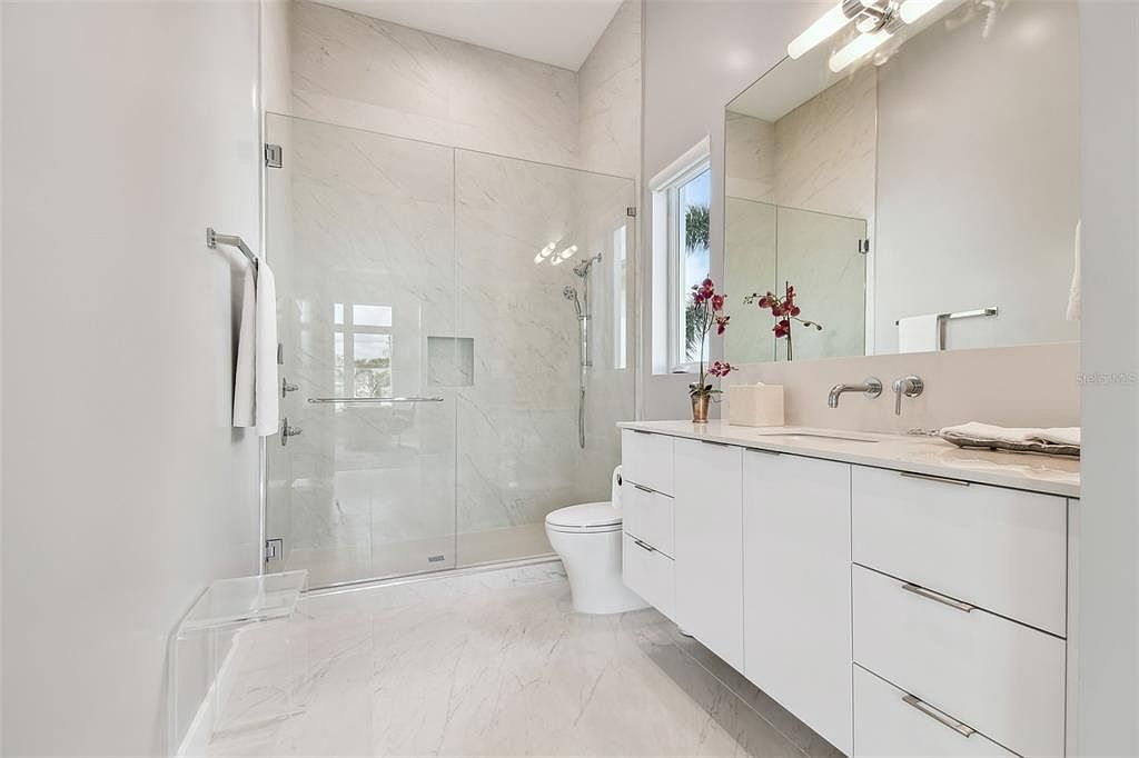 This is a bright and modern primary bathroom featuring a glass-enclosed shower with marble-like tile, a sleek white vanity with ample storage, and a large mirror. The room is well-lit and has a clean, minimalist aesthetic, creating a spa-like atmosphere. The flooring also appears to be marble or a similar material, adding to the luxurious feel.