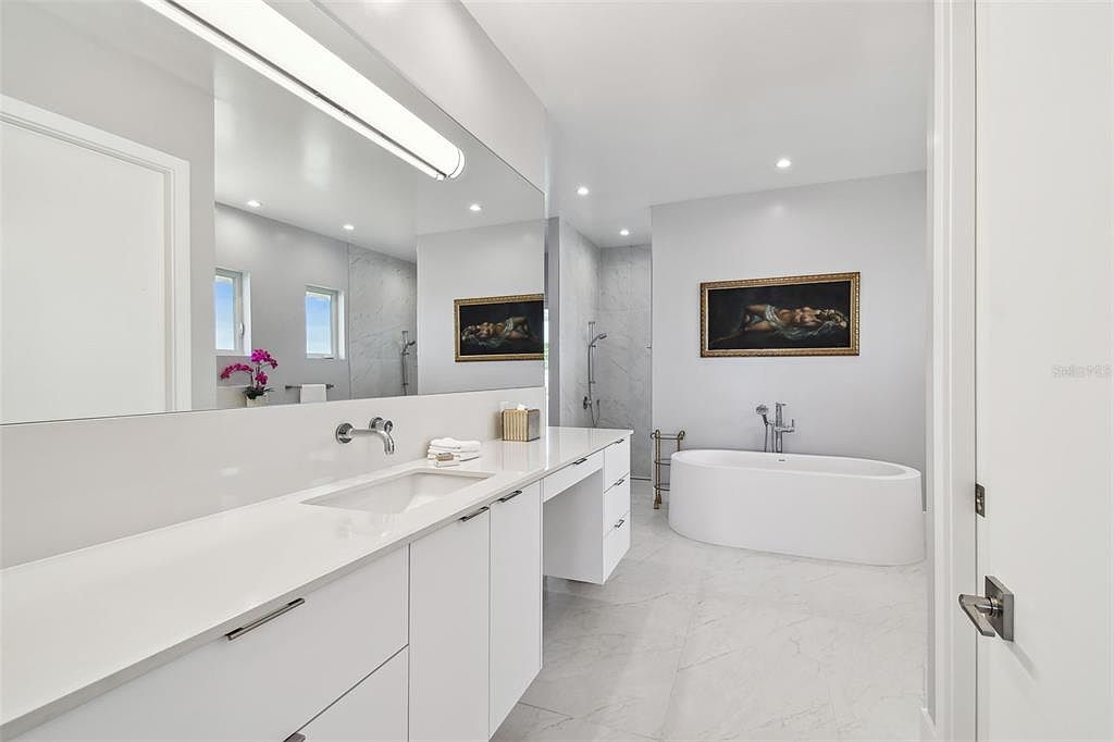 This is a bright and modern primary bathroom featuring a large vanity with white countertops and cabinets, a sleek undermount sink, and a large mirror with integrated lighting. A freestanding bathtub sits against a wall adorned with a framed artwork, and the floor is covered in light-colored marble tiles. The overall impression is clean, luxurious, and spa-like.