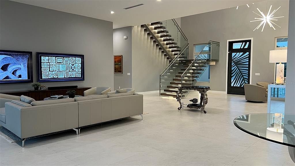 This is a spacious and modern living room featuring a large sectional sofa, contemporary artwork, and a striking staircase with glass railings. The room is well-lit with recessed lighting and a modern chandelier, and the light-colored tile flooring adds to the airy feel. The overall impression is one of luxury and sophistication.