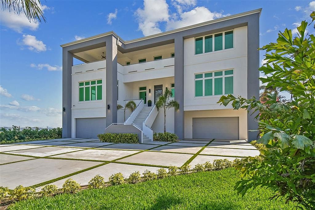 This is a front exterior view of a modern, multi-story home. The house features a gray and white color scheme, with large windows and a prominent staircase leading to the front entrance. The property includes a well-manicured lawn and a spacious driveway with grass accents, enhancing the curb appeal.