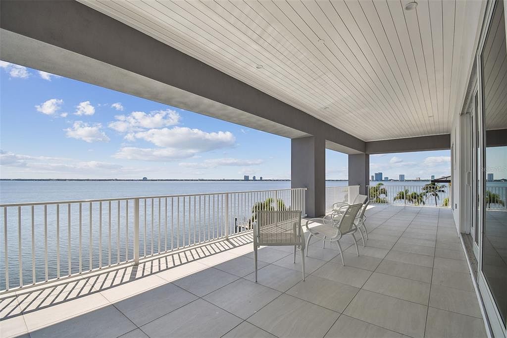 This image showcases a spacious balcony with a stunning waterfront view. The balcony features a white railing, gray tiled flooring, and several white chairs, creating a relaxing outdoor living space. The view includes a calm body of water and a distant cityscape, adding to the property's appeal.