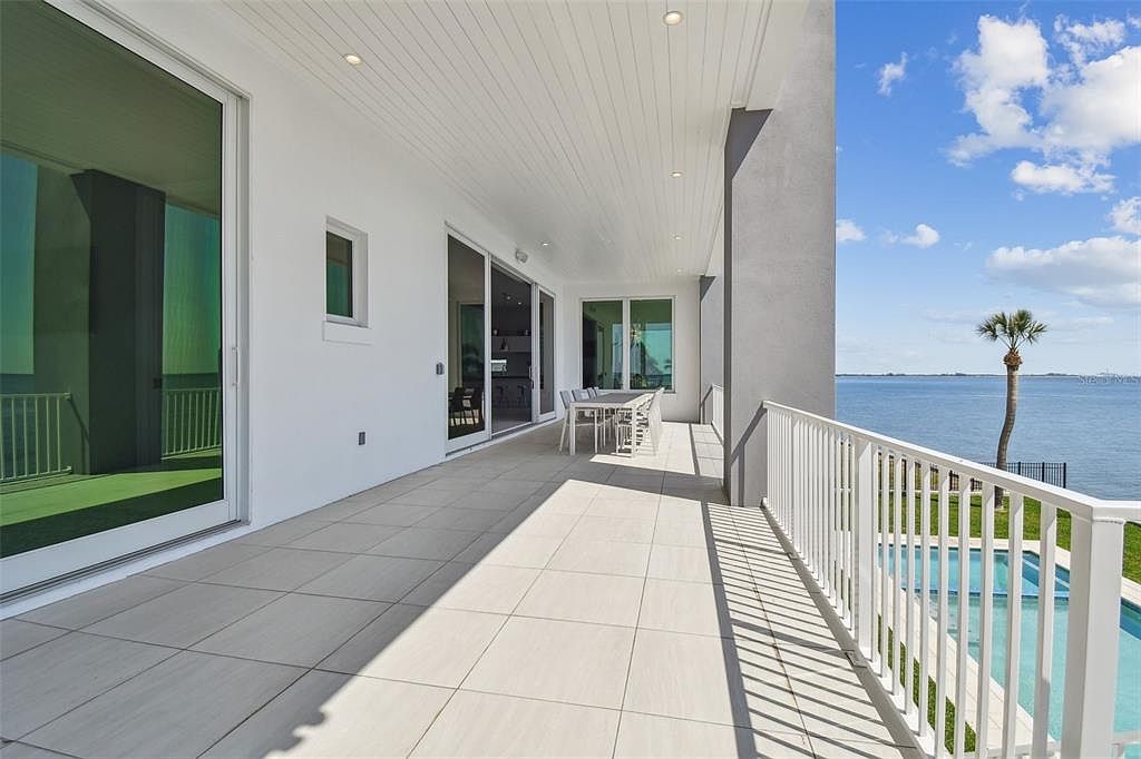 This is a view of a modern home's outdoor balcony or patio area. The space features light-colored tile flooring, a white railing, and a dining set, offering a relaxing outdoor living area with a view of the water and a palm tree. The design is clean and contemporary, emphasizing the connection between indoor and outdoor living.