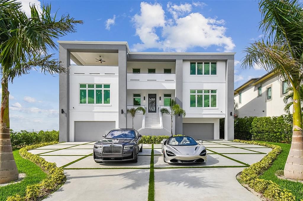 This is a front exterior view of a modern two-story home with a gray and white color scheme. The property features a well-manicured lawn, palm trees, and a driveway with two luxury cars parked in front. The architectural style is contemporary, emphasizing clean lines and a grand entrance with stairs leading to the front door.